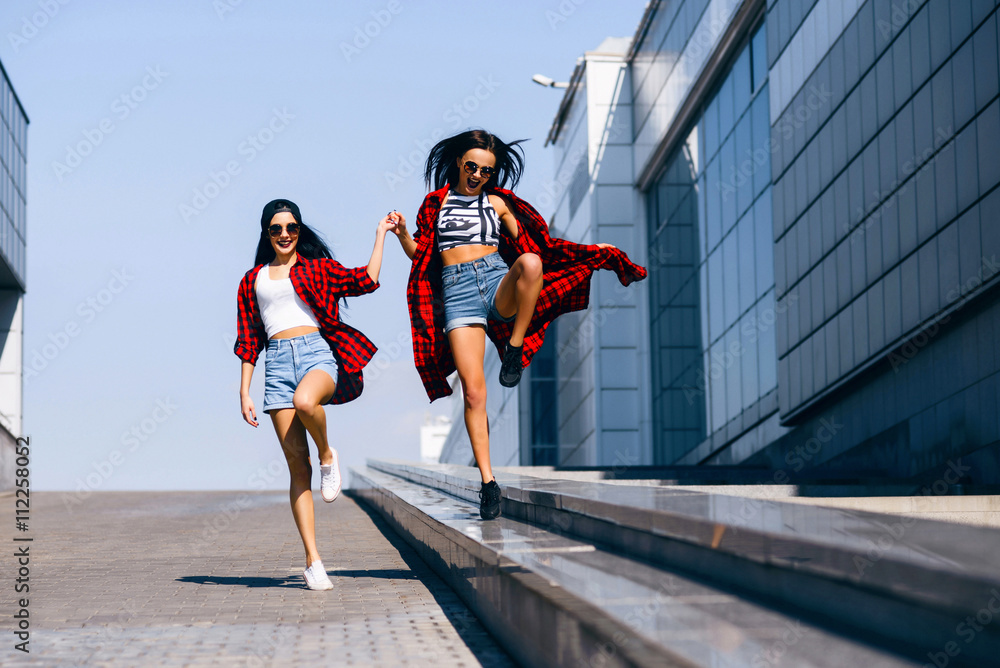 Two young happy hipster girls in sunglasses having fun, smiling, laughing, jumping, walking outdoor on the street, summer relax concept Two young happy hipster girls in sunglasses having fun, smiling, laughing, jumping, walking outdoor on the street, summer relax concept