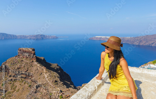 Attractive woman wearing hat and yellow dress enjoying the view of volcanic island in the early morning on Santorini, Mediterranean sea, Greece