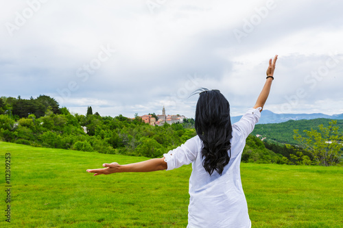 Back of woman with black hair raising hands and blurred ancient buildings and church on top of hill surrounded by trees on cloudy day, Labin, Croatia 