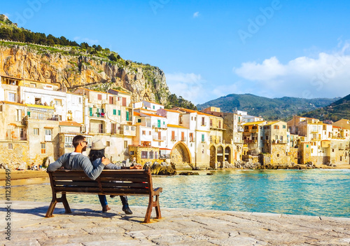 Couple sitting on bench, enjoying view of beach town of Cefalu in Sicily, Italy