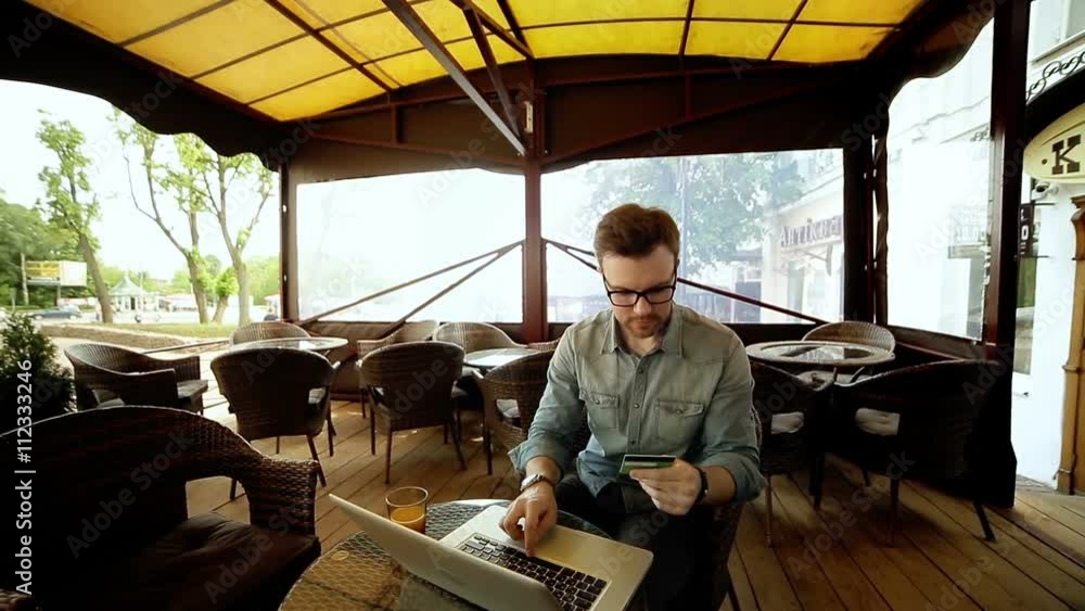 Handsome man in shirt and glasses using a laptop at cafe while holding credit card in the hands, on-line shopping outdoors. Copy space Handsome man in shirt and glasses using a laptop at cafe while holding credit card in the hands, on-line shopping outdoors. Copy space