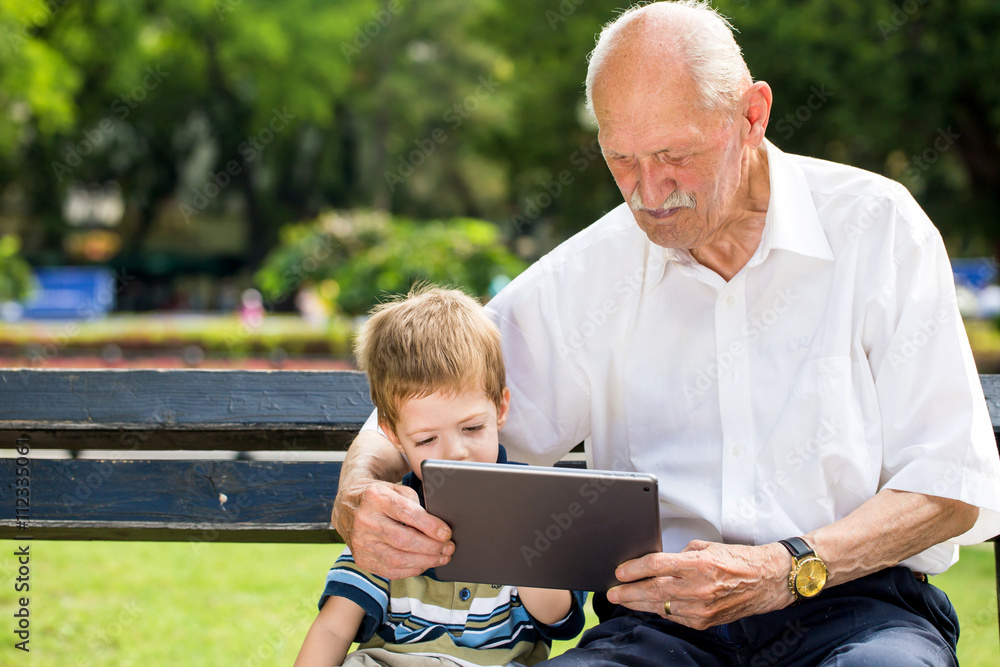 Grandchild teaching to his grandfather to use tablet on a bench in summer sunny day Grandchild teaching to his grandfather to use tablet on a bench in summer sunny day