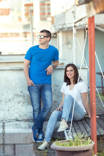 Young couple is relaxing outdoors. Young woman is sitting on wooden swing and looking at camera. Young man is standing next to his girfriend and looking away.