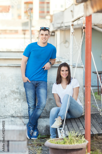 Young couple is relaxing outdoors. Young woman is sitting on wooden swing and young man is standing next to her. They are looking at camera and smiling.