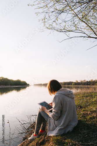Young girl works with the tablet on the banks of the river