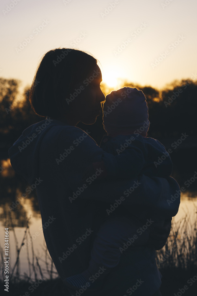 Young mother holds her child at the hands of on the banks of the river at sunset