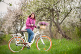 Active female biker wearing purple jacket and jeans starting to ride a vintage white bicycle and lilac flowers basket, against the background of blooming trees in spring garden