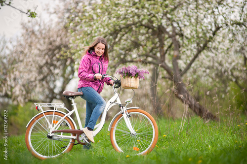 Active female biker wearing purple jacket and jeans starting to ride a vintage white bicycle and lilac flowers basket, against the background of blooming trees in spring garden