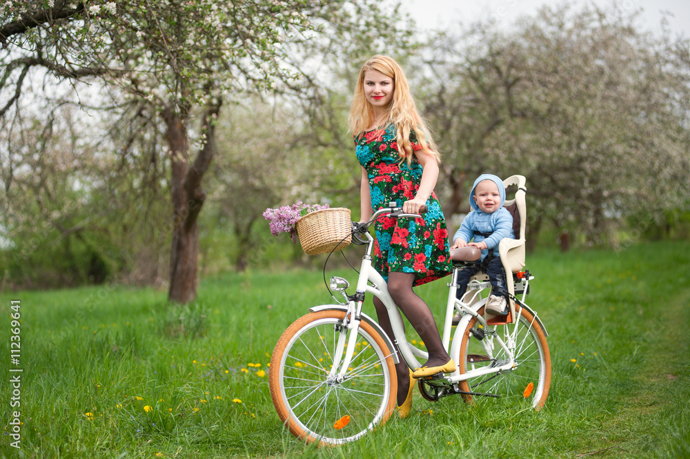 Mother with long blonde hair in dress riding bicycle with baby in bicycle chair. A bouquet of lilacs lay in the basket. Blooming fresh greenery in spring garden on the background