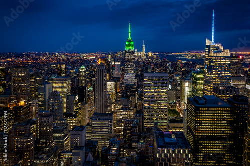 Aerial view of midtown Manhattan at night, the heart of a financial empire that dominates the business world, with every building vividly colored in shades of blue and green in NYC