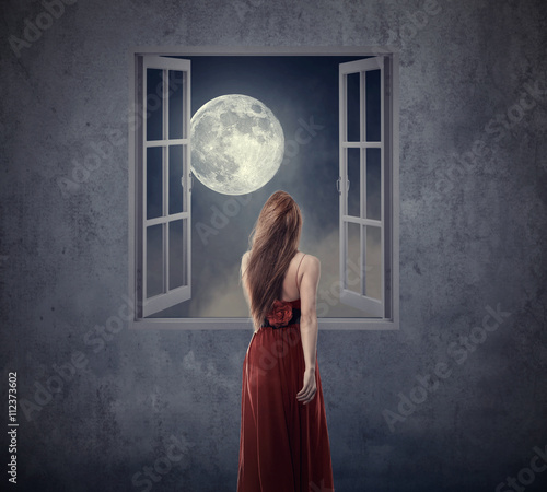 Fotografie Beautiful woman in red dress walking to opened window with moon