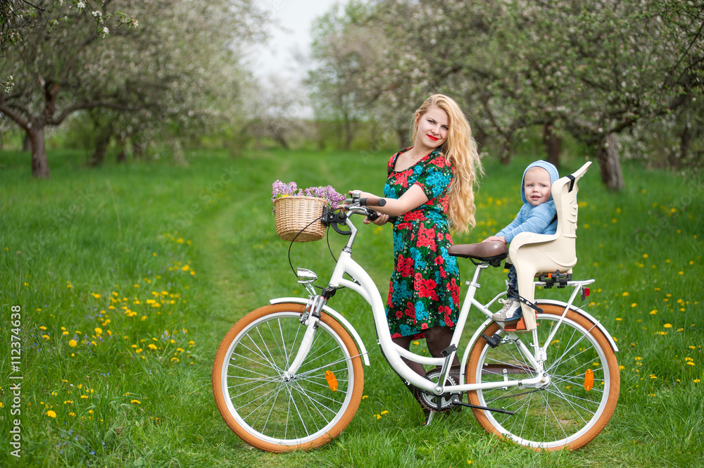 Smiling mother in flowered dress holding bike with child who sits in bicycle chair, against the background of blooming trees, dandelions and fresh greenery in spring garden
