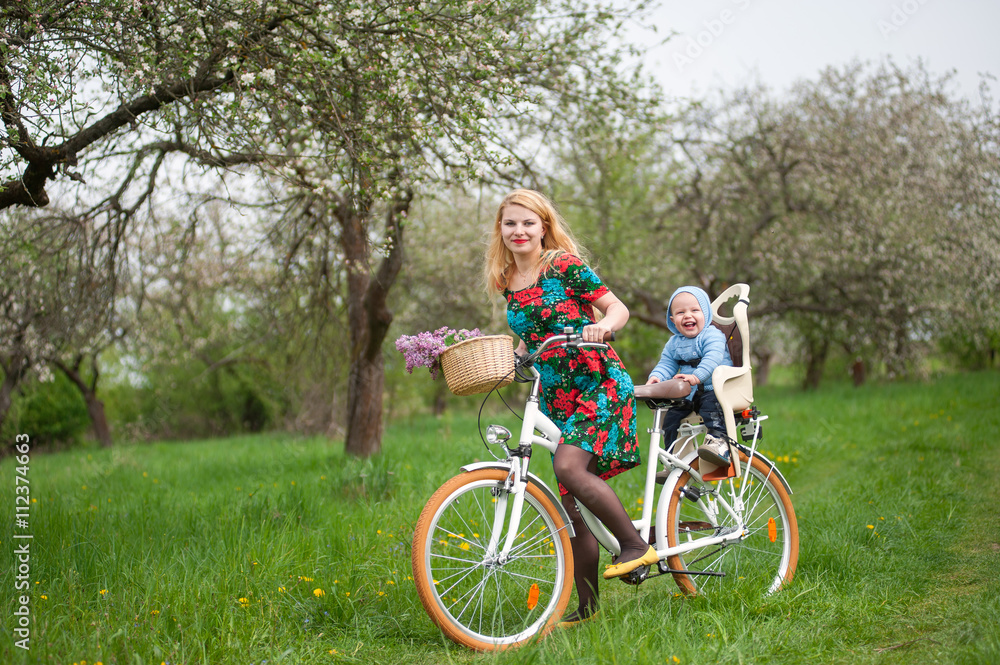 Mother with long blonde hair in dress riding city bike with baby in bicycle chair, in the basket lay a bouquet of lilacs, against the background of blooming fresh greenery in spring garden Mother with long blonde hair in dress riding city bike with baby in bicycle chair, in the basket lay a bouquet of lilacs, against the background of blooming fresh greenery in spring garden