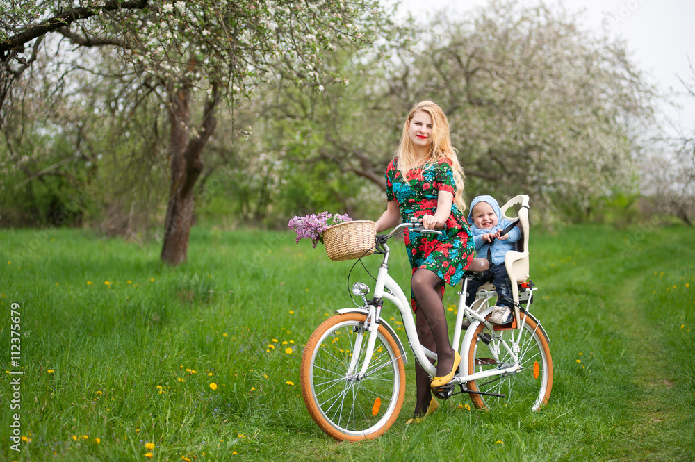 Naklejka premium Mother with long blonde hair in dress keeps bicycle and baby in bicycle chair, in the basket lay a bouquet of lilacs, against of blooming fresh greenery in spring garden on the background