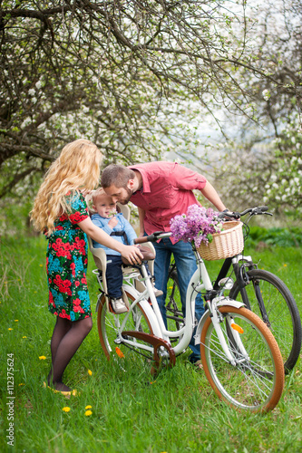 Young family on a bicycles in the spring garden. Mother holding your bike and baby sitting in bicycle chair, in the basket lay a bouquet of lilacs, against the background of blooming fresh greenery
