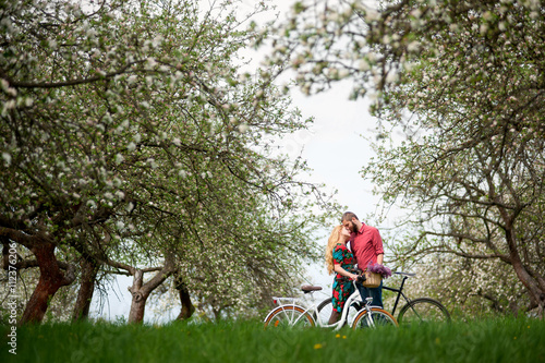 Young couple of bikers loving and keeping bikes against the background of blooming trees and fresh greenery in spring garden. Couple together enjoying romantic holidays. Side view