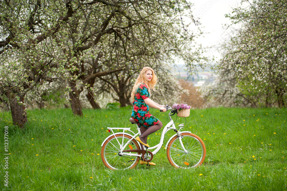 Young female with long blond hair wearing flowered dress and yellow shoes riding a vintage white bicycle with flowers in basket. Blooming trees, dandelions and greenery in spring garden. Side view