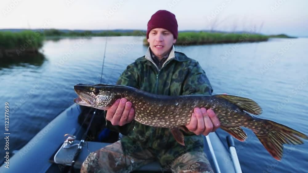 Young fisherman holding pike and sitting in the boat on the lake Young fisherman holding pike and sitting in the boat on the lake