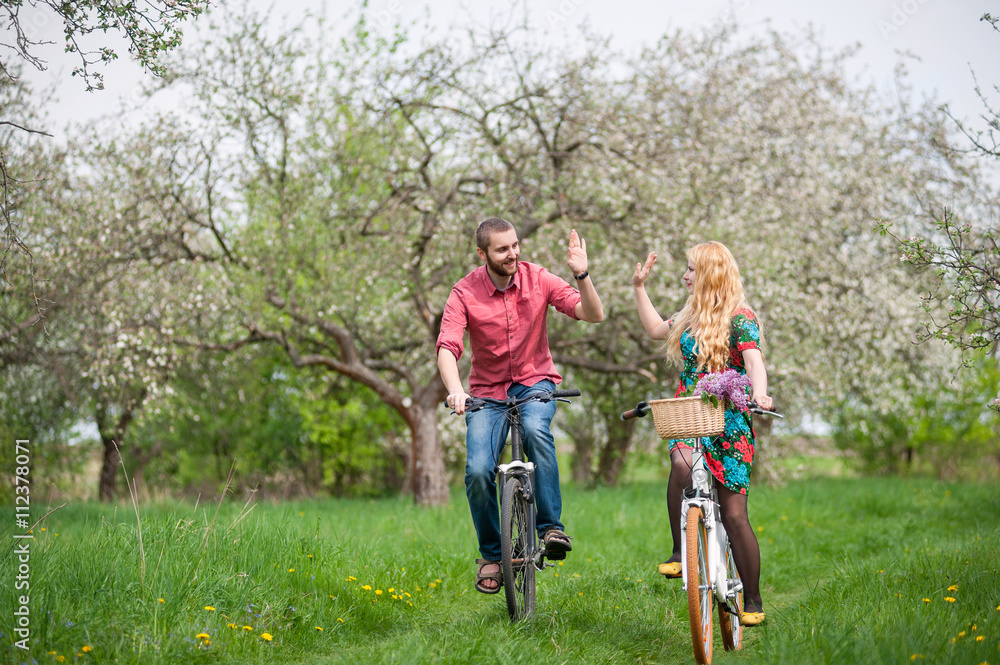 Successful couple riding bicycles in the blooming spring garden and giving each other a high five. Woman with long blond hair wearing flowered dress and man in a red shirt and jeans Successful couple riding bicycles in the blooming spring garden and giving each other a high five. Woman with long blond hair wearing flowered dress and man in a red shirt and jeans