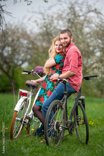 Two smiling lovers standing with bicycles and looking to the camera in the spring garden, against the background of trees and fresh greenery