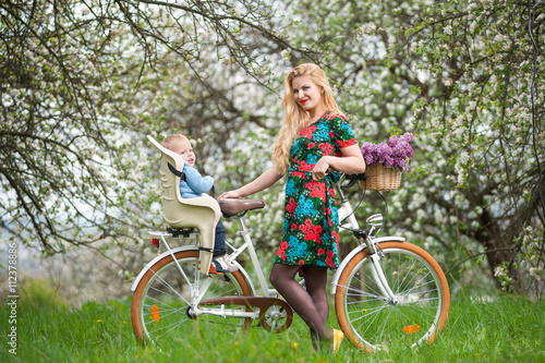 Fototapeta Naklejka Na Ścianę i Meble -  Smiling blonde female with long hair and red lips in flowered dress holding bike and baby in bicycle chair, in the basket lay a bouquet of lilacs. Blooming trees in spring garden on the background