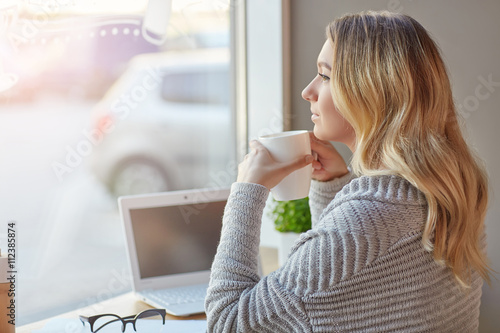 Beautiful young woman working with computer at cafe holding a cup of drink and looking thoughtfully out the window . Freelancer