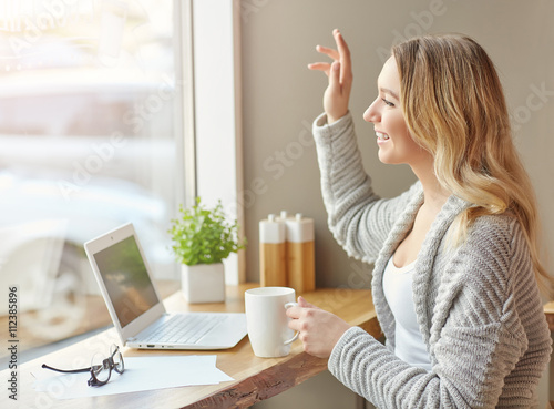 Hi. Beautiful young woman working with computer at cafe holding a cup of drink and waving in the window.