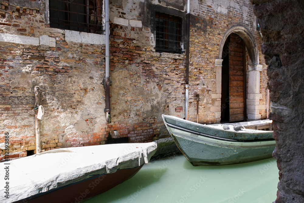 Two boats in the narrow channel of water a milky green. Old brick wall with crumbling plaster, arched doorway.