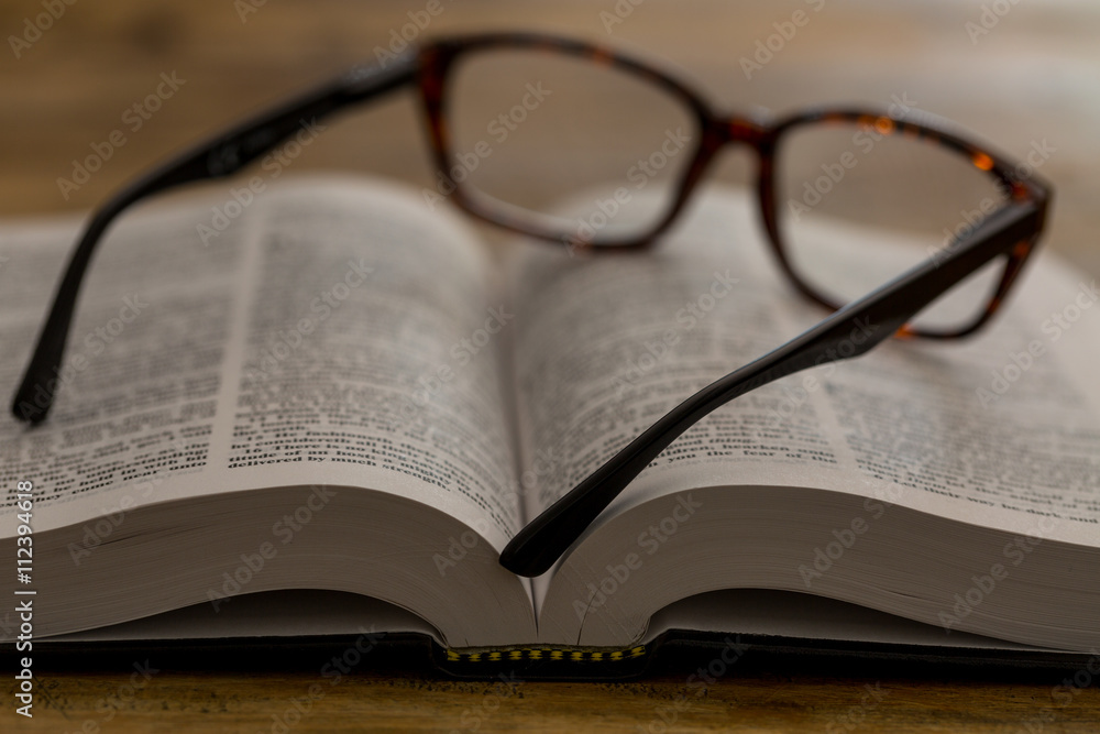 Aufgeschlagenes Buch mit Lesebrille auf dunklem Holztisch, Nahan Aufgeschlagenes Buch mit Lesebrille auf dunklem Holztisch, Nahan