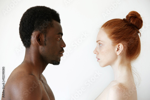 Multi-ethnic relationships concept. Headshot of shirtless African man and freckled redhead woman looking at each other with loving and caring expression posing against white concrete wall background