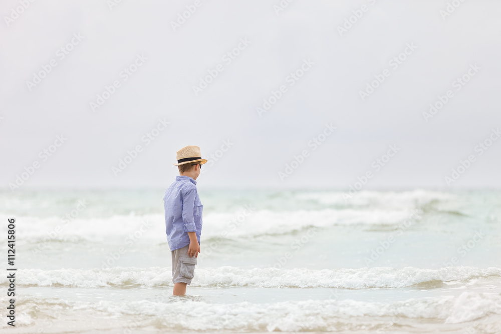 Portrait of adorable kid boy in straw hat and sun glasses walking on ocean beach and looking at the waves. Vacations by the sea. Outdoor.