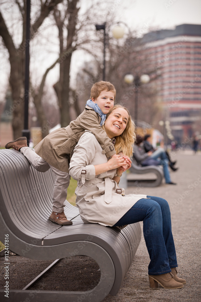 Beautiful young mother hugging her adorable kid boy in the park on a spring day. Happy family in trench coats walking outdoors and having fun. Mother's love. Beautiful young mother hugging her adorable kid boy in the park on a spring day. Happy family in trench coats walking outdoors and having fun. Mother's love.
