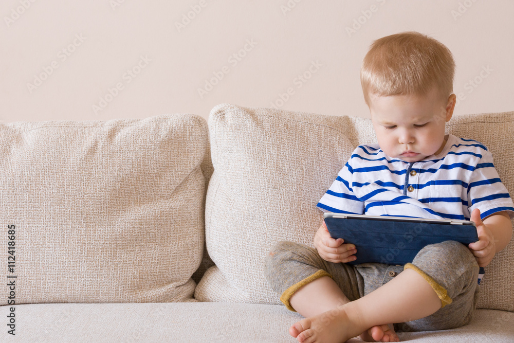Adorable blond toddler boy laying on the sofa and playing with tablet pc at home, indoors. Child with tablet computer. Adorable blond toddler boy laying on the sofa and playing with tablet pc at home, indoors. Child with tablet computer.