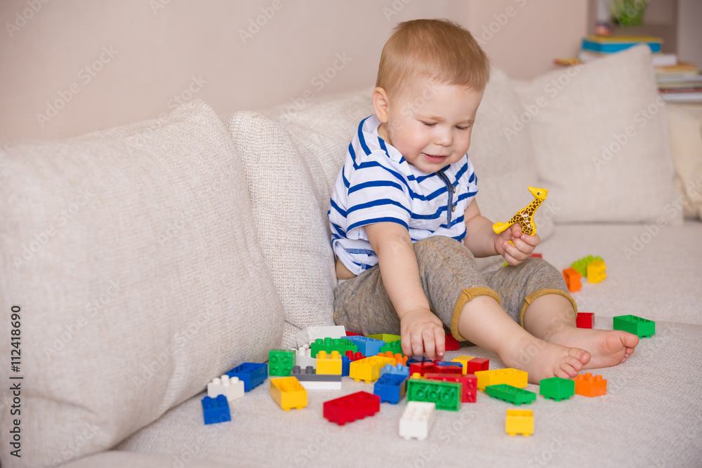 Cute little toddler boy in a striped shirt playing colorful plastic blocks on the sofa indoors. child having fun and building out of bright constructor bricks. Early learning. Creative. Cute little toddler boy in a striped shirt playing colorful plastic blocks on the sofa indoors. child having fun and building out of bright constructor bricks. Early learning. Creative.