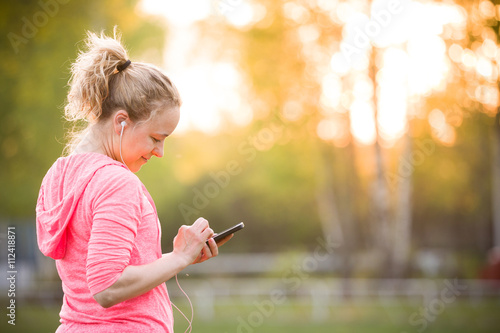 Young beautiful woman choosing music for her workout in the park on a sunny summer day. Sportive girl jogging on the stadium. Healthy lifestyle and sport concept.