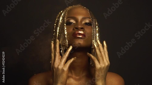 Surreal Portrait of Sexy African American Woman with Glossy Golden Makeup and Headwear Posing on Black Studio Background. Bronze Bodypaint