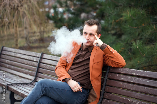 A young man sits on a bench and smokes an electronic cigarette. © davit85
