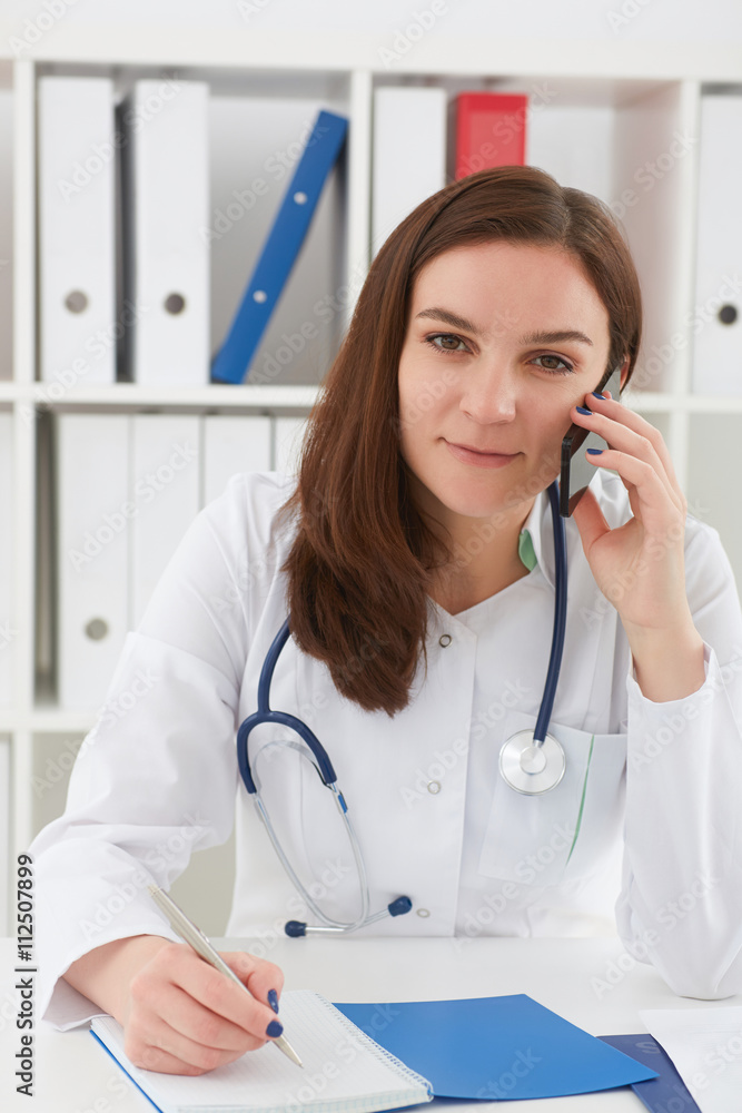 Portrait of a beautiful medical doctor talking on the phone and writing something in a notebook. Portrait of a beautiful medical doctor talking on the phone and writing something in a notebook.