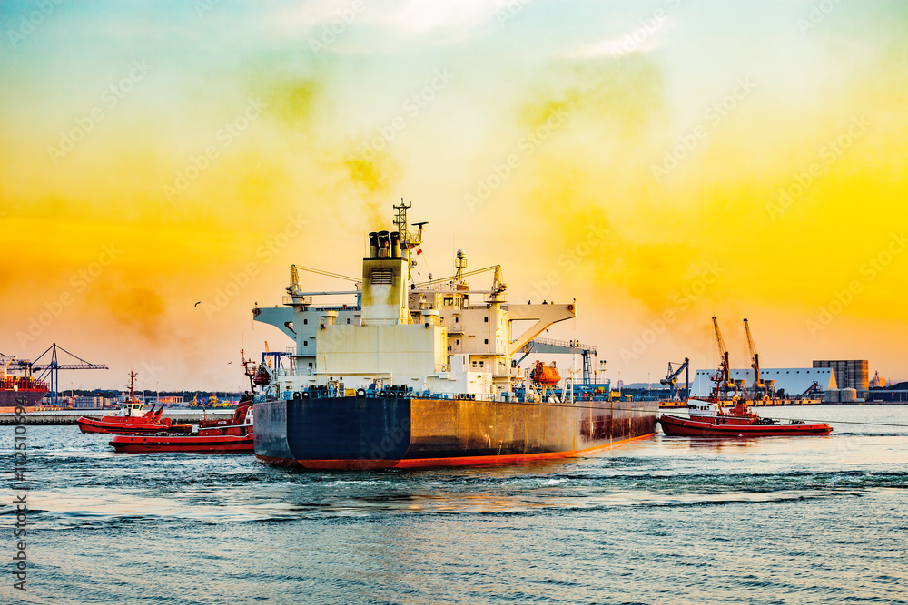 Fototapeta premium Big ship with escorting tugs leaving port at sunset.