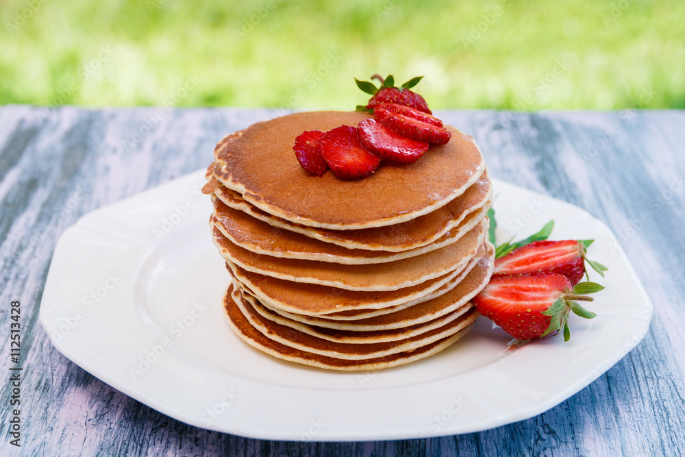 Pancakes with fresh strawberry and mint on white plate on pink wooden background  in garden or on nature background.  Stack of pancakes on table.