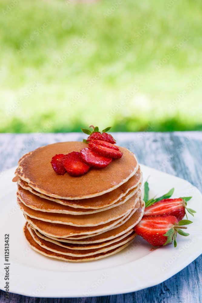 Pancakes with fresh strawberry and mint on white plate on pink wooden background in garden or on nature background. Stack of pancakes on table. Pancakes with fresh strawberry and mint on white plate on pink wooden background in garden or on nature background. Stack of pancakes on table.