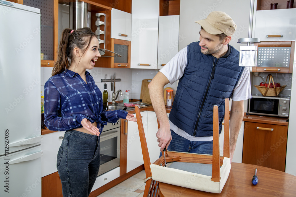 Handyman repairing furniture in the kitchen. He repairs a chair with a screwdriver. The man looks at a housewife and smiling. The woman looks at him, smiles and shrugs. She wonders.