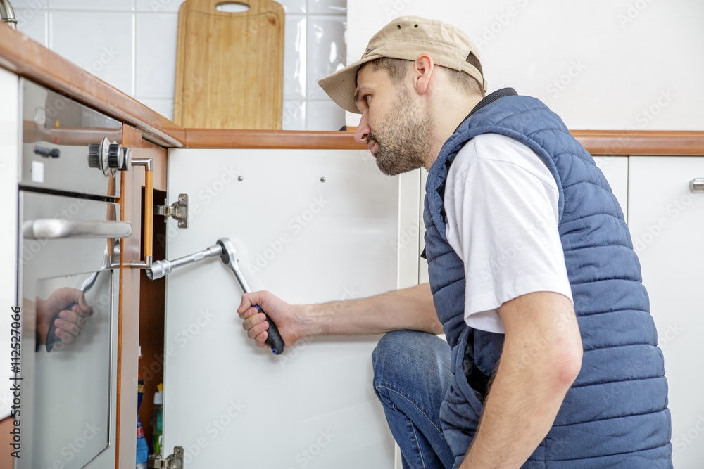 Worker repairing the sink in the kitchen. Man looks at the sink and holding a wrench.