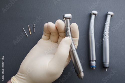 Hand of dentist in latex protective gloves holding dental drill handpiece. Flat lay of dental handpiece in dentist hand on gray background. Dental handpieces and bur tools on background photo