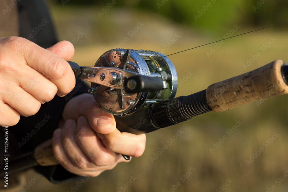 Man fishing with reel and rod. One hand on the crank and reeling fishing  line in to the round reel and other hand holding fishing rod with blurred  nature background. Stock Photo