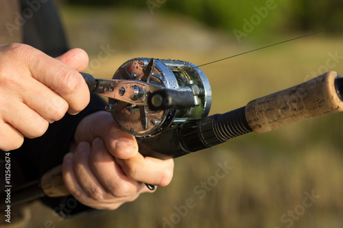 Man fishing with reel and rod. One hand on the crank and reeling fishing line in to the round reel and other hand holding fishing rod with blurred nature background. 