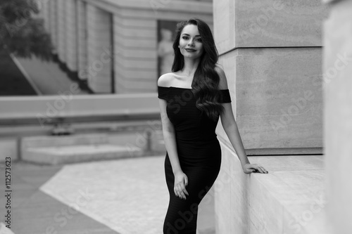 Black and white portrait of young beautiful elegant woman in black dress. Pretty sensual girl with long curly hair posing outdoors at city street