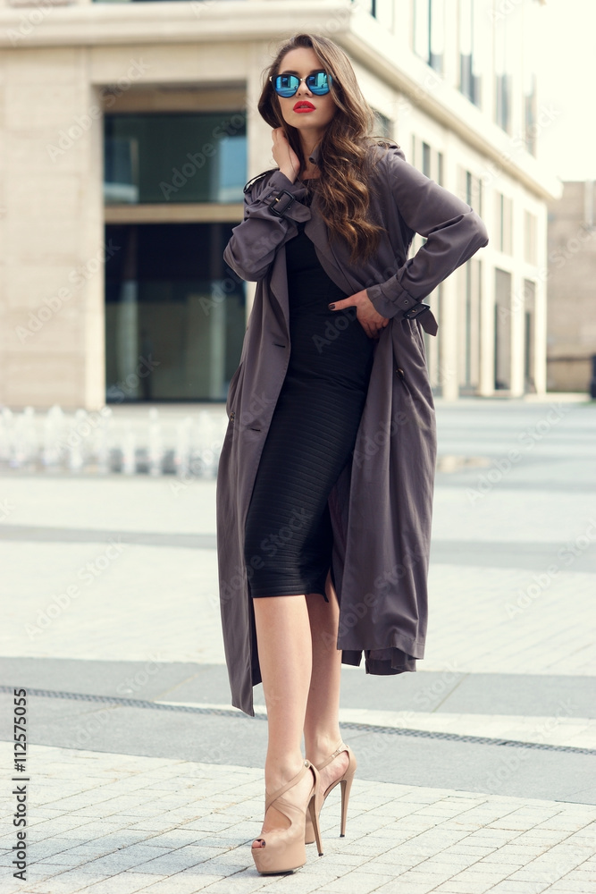 Outdoor dynamic fashion portrait of young beautiful stylish woman in black dress, grey coat and sunglasses walking on a windy day against city background