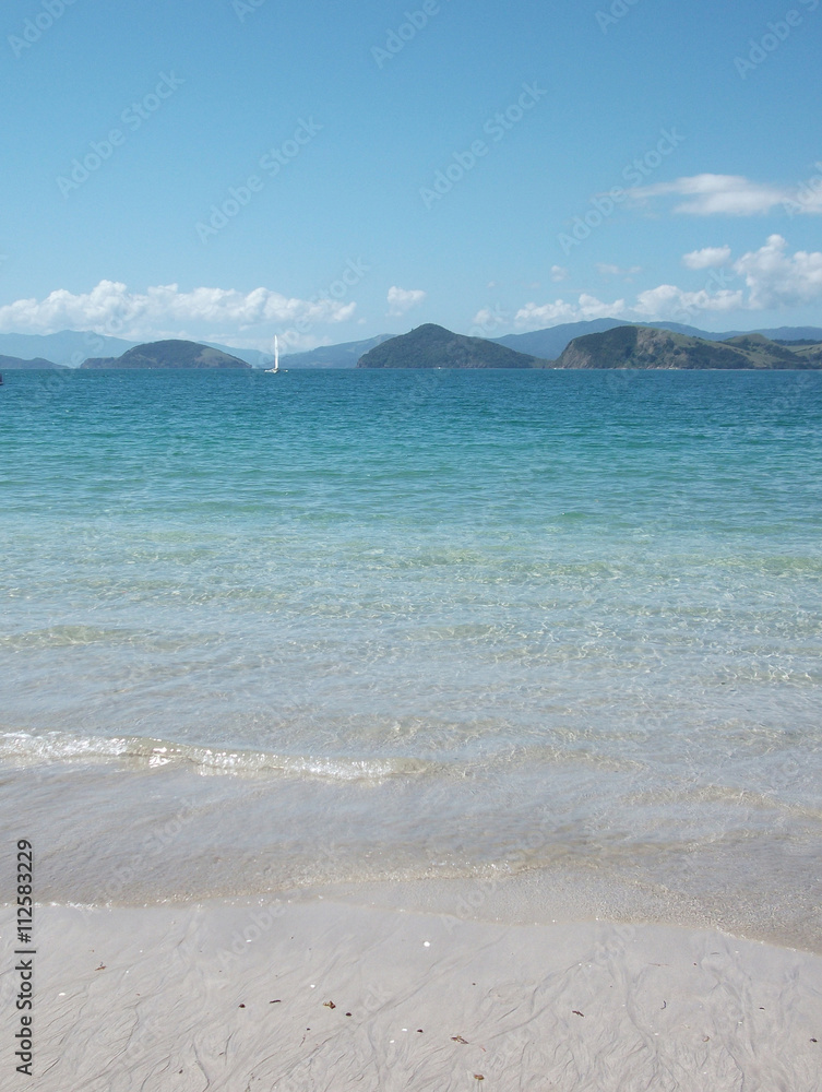 Clear blue sea, blue sky and white sand beach near Te Kouma - Coromandel Peninsula, New Zealand. A sailboat is in the distance.