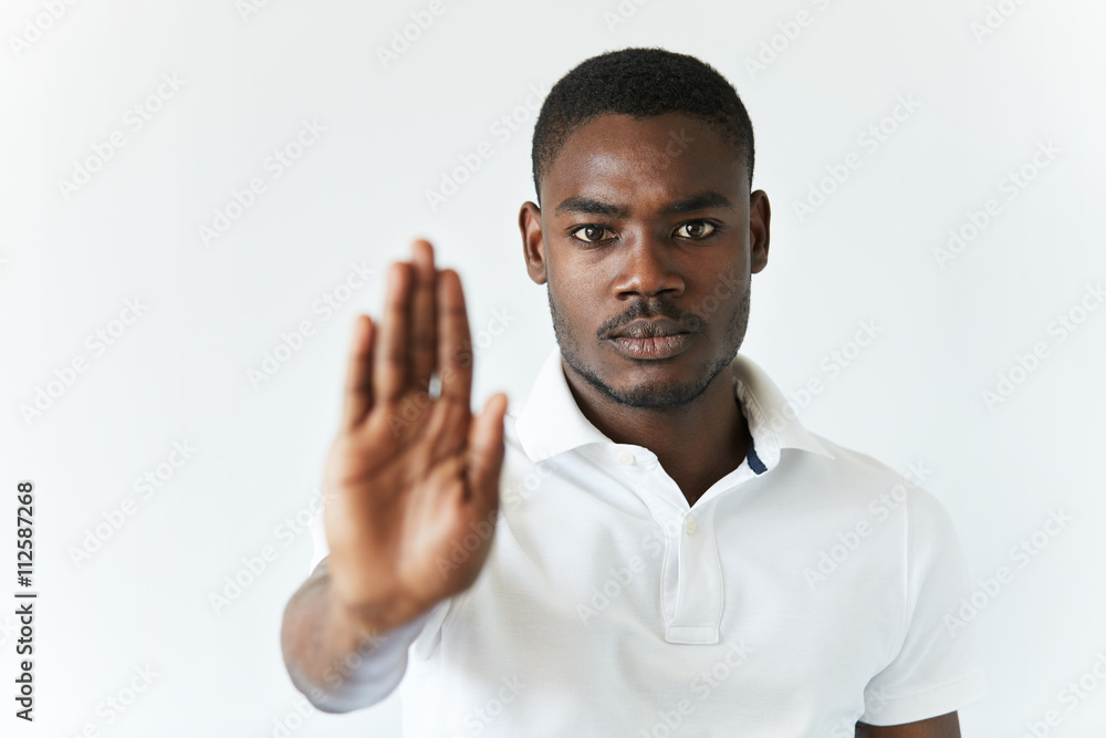 Headshot of dark-skinned young man wearing white polo shirt, preventing you from doing something with his open palm, looking at the camera with serious expression. Selective focus on the face Headshot of dark-skinned young man wearing white polo shirt, preventing you from doing something with his open palm, looking at the camera with serious expression. Selective focus on the face